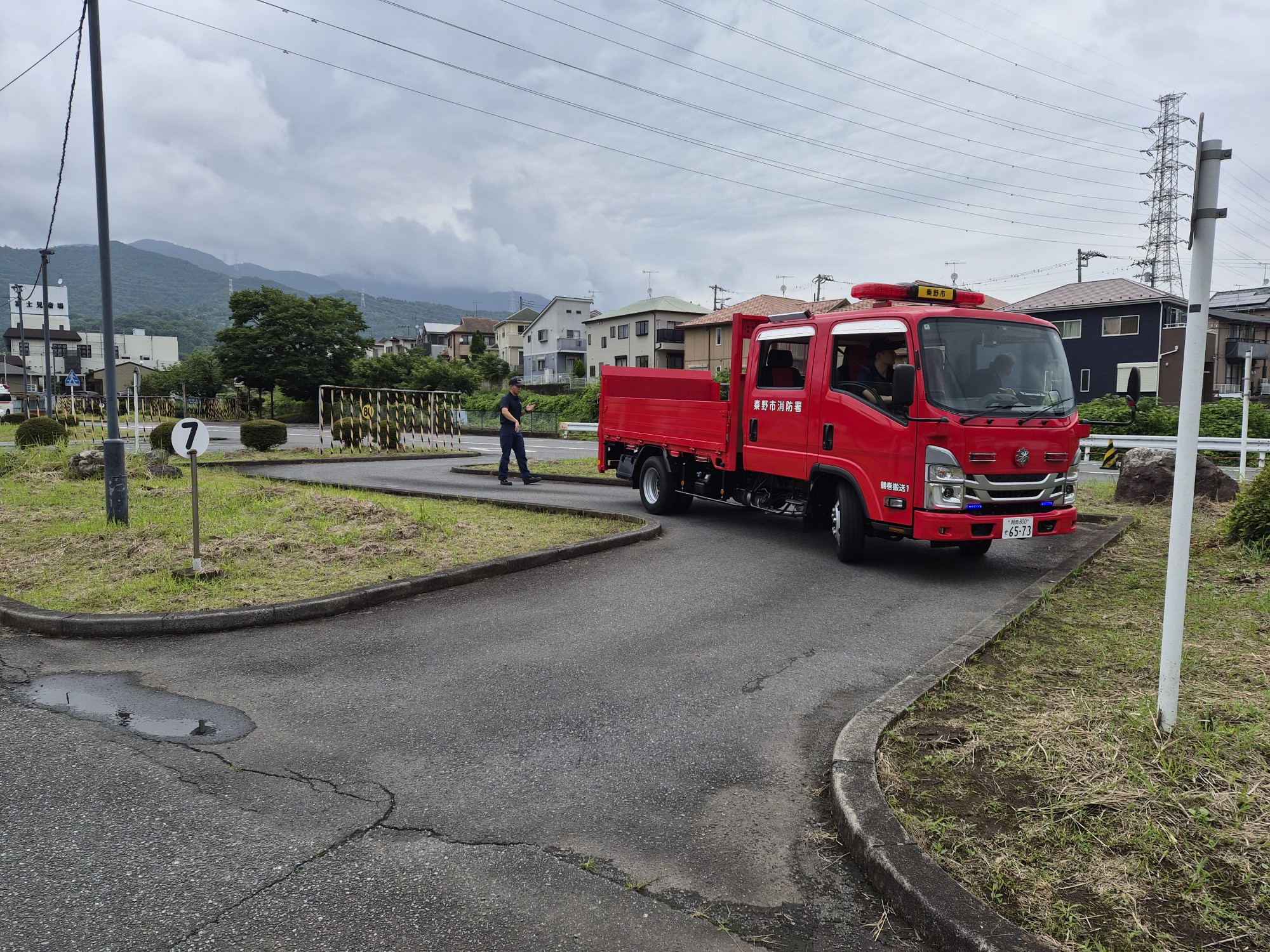 教習コースの狭いカーブを秦野市消防本部の赤いトラックがクランク走行し、その様子を隊員が見守っている写真