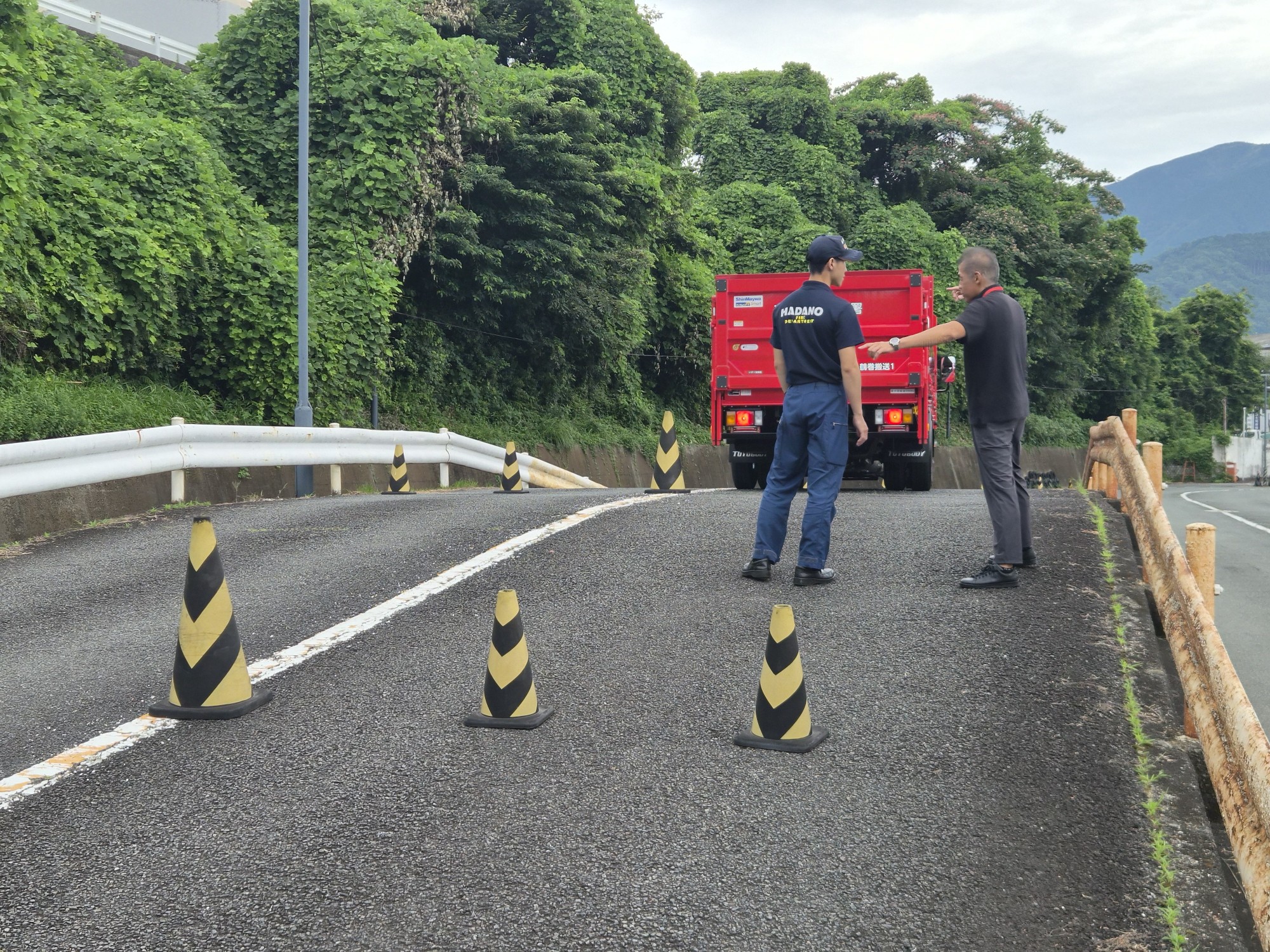 緑豊かな山道を背景に、秦野市消防本部の赤い消防車が停車し、隊員たちがコーンを置いて活動準備の指導を受けている様子の写真