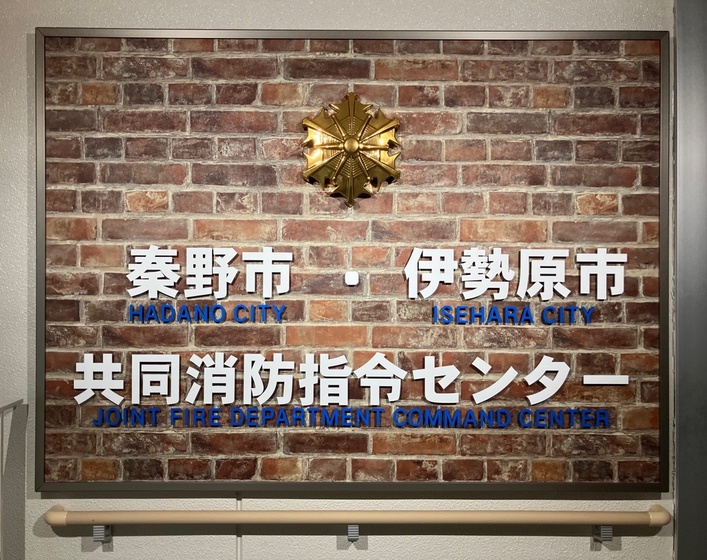 レンガ調の壁に、秦野市と伊勢原市の共同消防指令センターであることを示す文字が日本語と英語で大きく記載されており、中央上部には金色に輝く消防の紋章が取り付けられている看板の写真