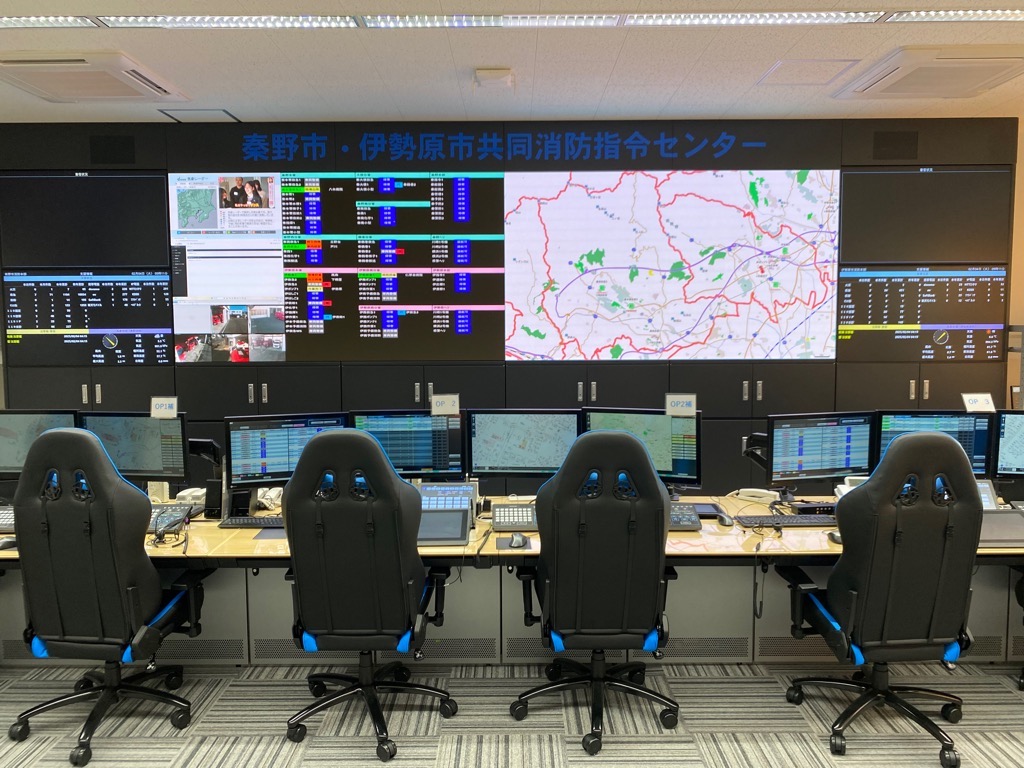 手前には4脚の黒と青のゲーミングチェアが並び、奥の壁には「秦野市・伊勢原市共同消防指令センター」の文字と地図や運行状況などが表示された巨大なマルチディスプレイが設置されている写真