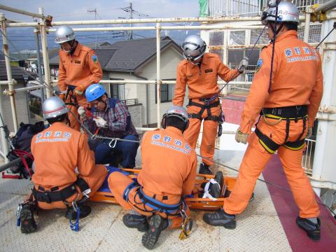 オレンジ色の救助服とヘルメットを着用した複数の救助隊員と山岳遭難救助隊が、地上からやや高い足場の上で、負傷者役の人をストレッチャーに収容する訓練を行っている様子の写真