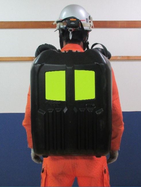 オレンジ色の救助活動服を着た隊員が、背中の黒い空気呼吸器の容器に2枚の大きな蛍光イエローの反射材が取り付けられている状態で立っている後姿の写真