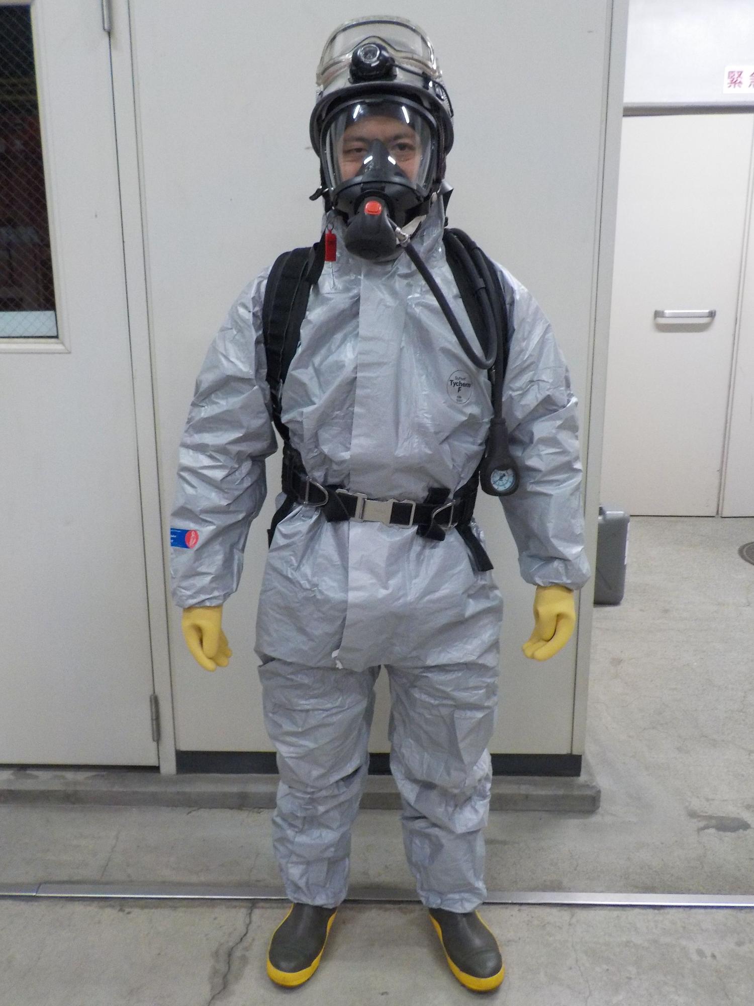 薄い灰色の化学防護服と黄色の手袋、黒と黄色の長靴を着用し、ヘルメットとフルフェイスマスクの空気呼吸器を装着した隊員が、建物内で正面を向いて立っている写真