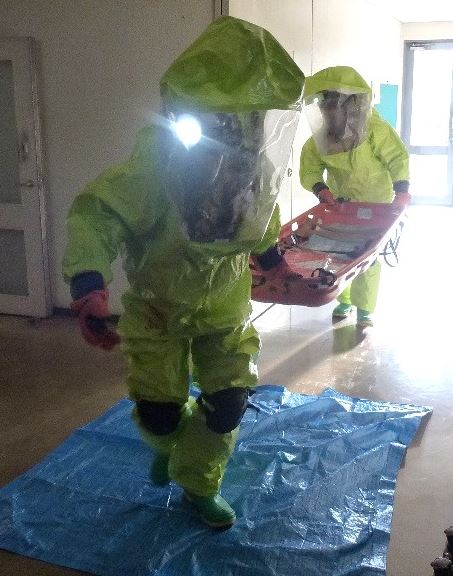 室内で緑色の化学防護服を着用した2人の隊員が、青いシートの上で、要救助者を搬送するためのストレッチャーを運んでいる、災害対応訓練の様子の写真