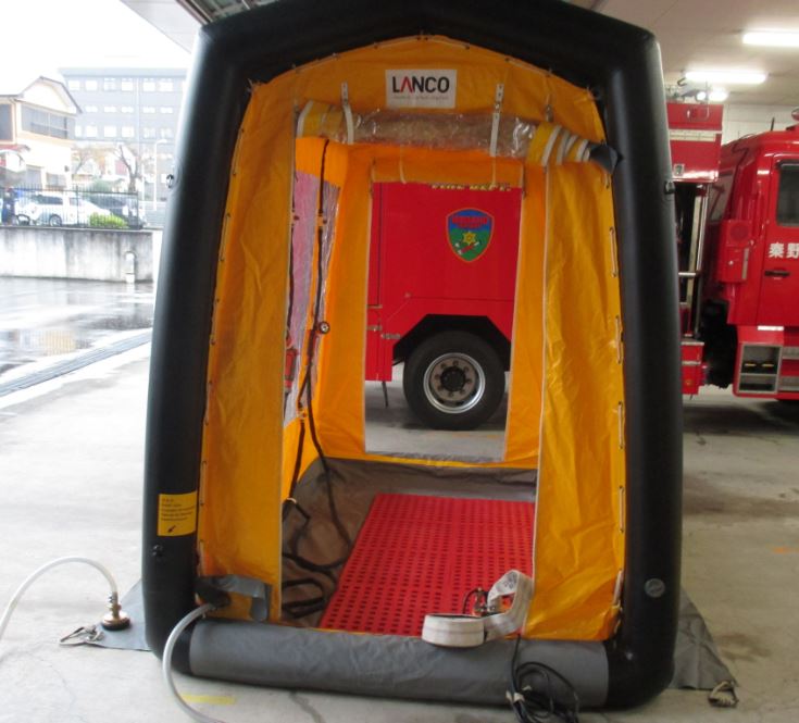 消防署の車庫内で、黒いフレームと黄色い内装の空気注入式除染テントが設置されており、テントの奥には赤い消防車の車体とタイヤの一部が見えている写真