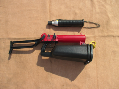 黒い伸縮式ストックと赤い本体、黒いグリップと弾倉状の部分からなる銃器のような形状をした救助用資機材と、その上に置かれた円筒形の黒いカートリッジの写真