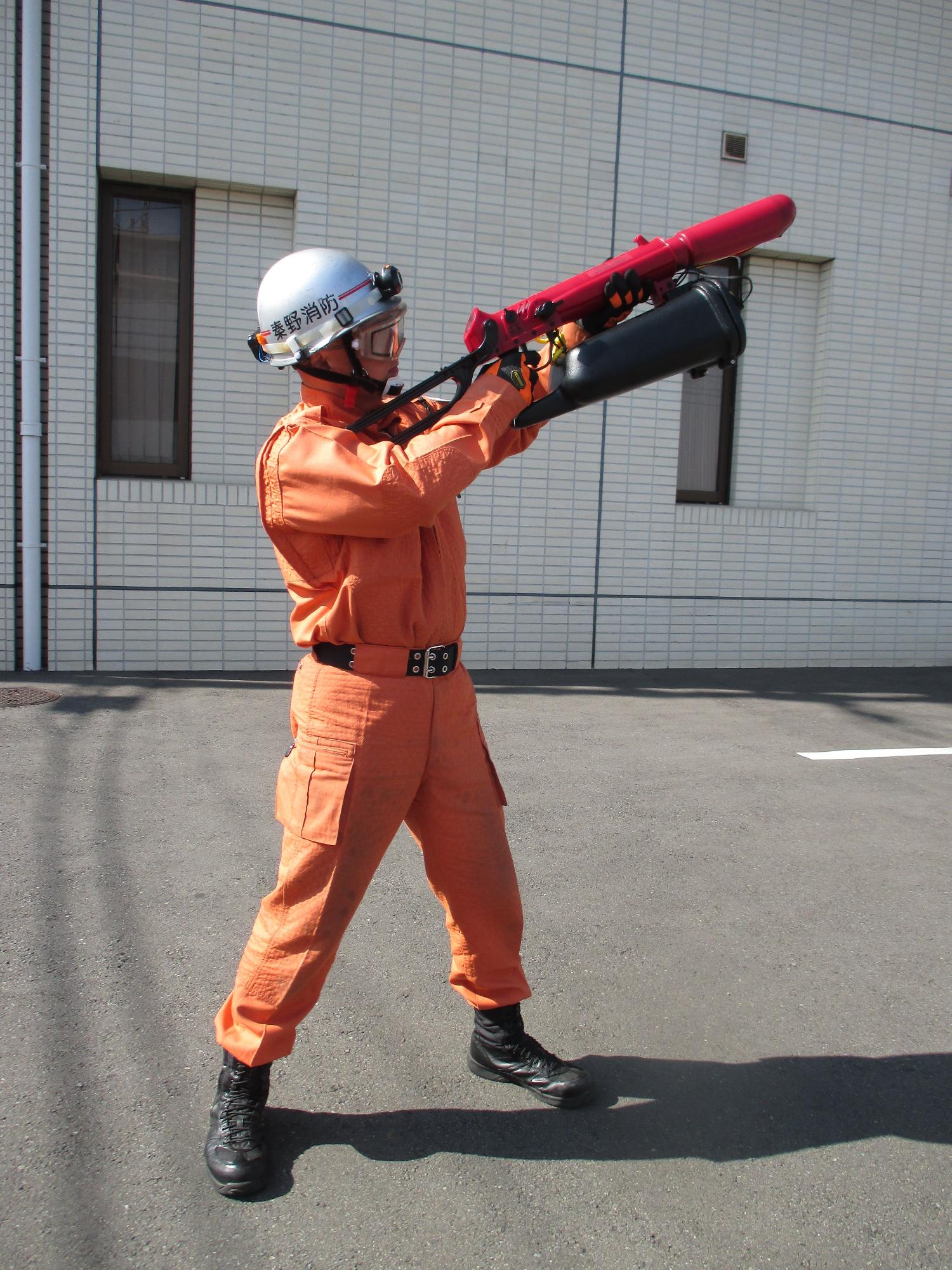 オレンジ色の活動服とヘルメットを着用した隊員が、肩に赤い発射筒と黒い弾倉状の部品からなる銃器のような救助用資機材を構えている全身写真