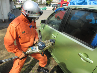 秦野消防のヘルメットを装着したオレンジ色の救助活動服の隊員が、緑色の乗用車のドアに油圧式救助器具のスプレッダーを差し込み、車体を開放する訓練を行っている様子の写真