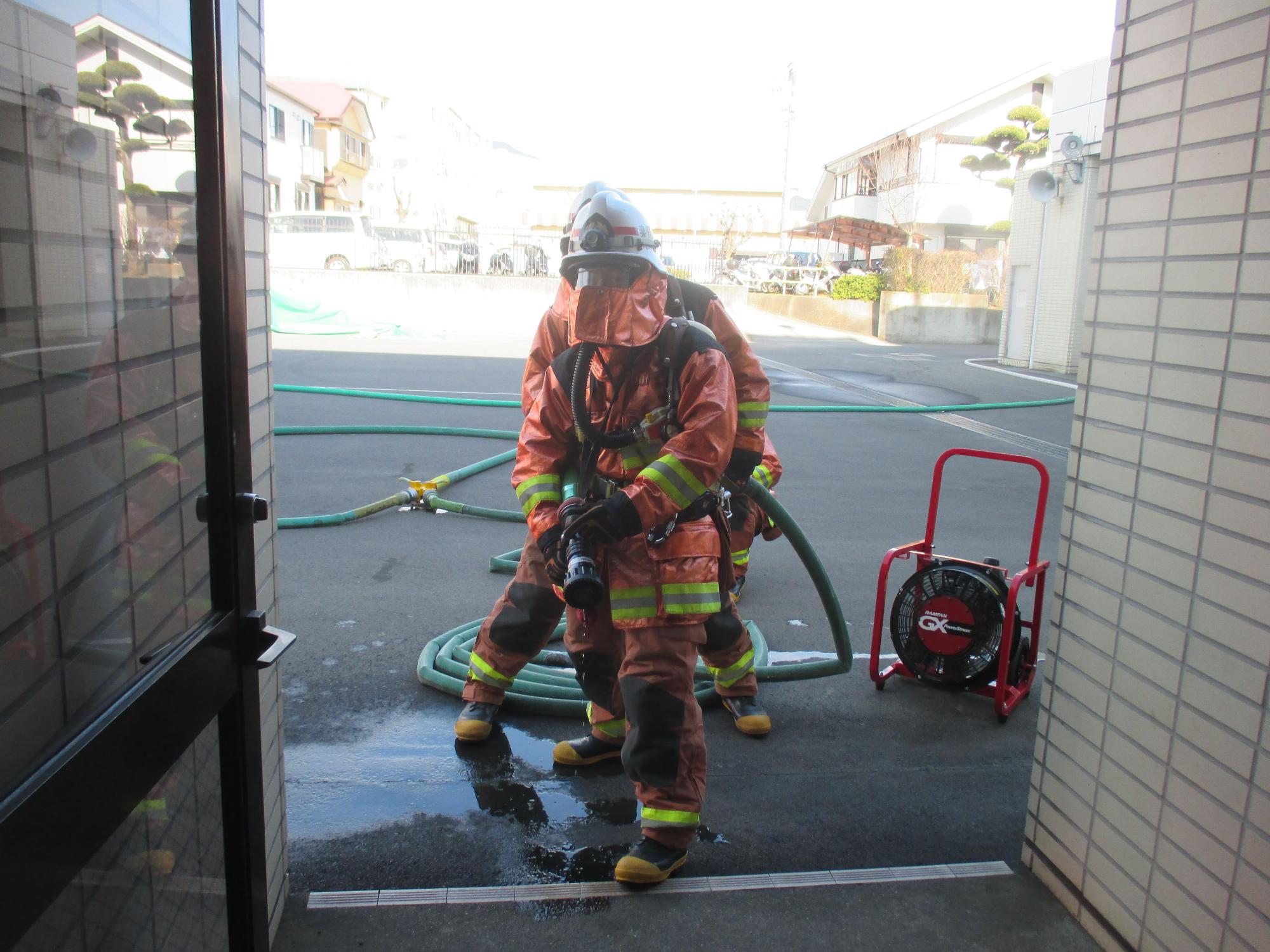 建物の開口部から明るい屋外を見ており、オレンジ色の防火服と空気呼吸器を装着した2人の消防士がホースとノズルを持って待機している様子の写真