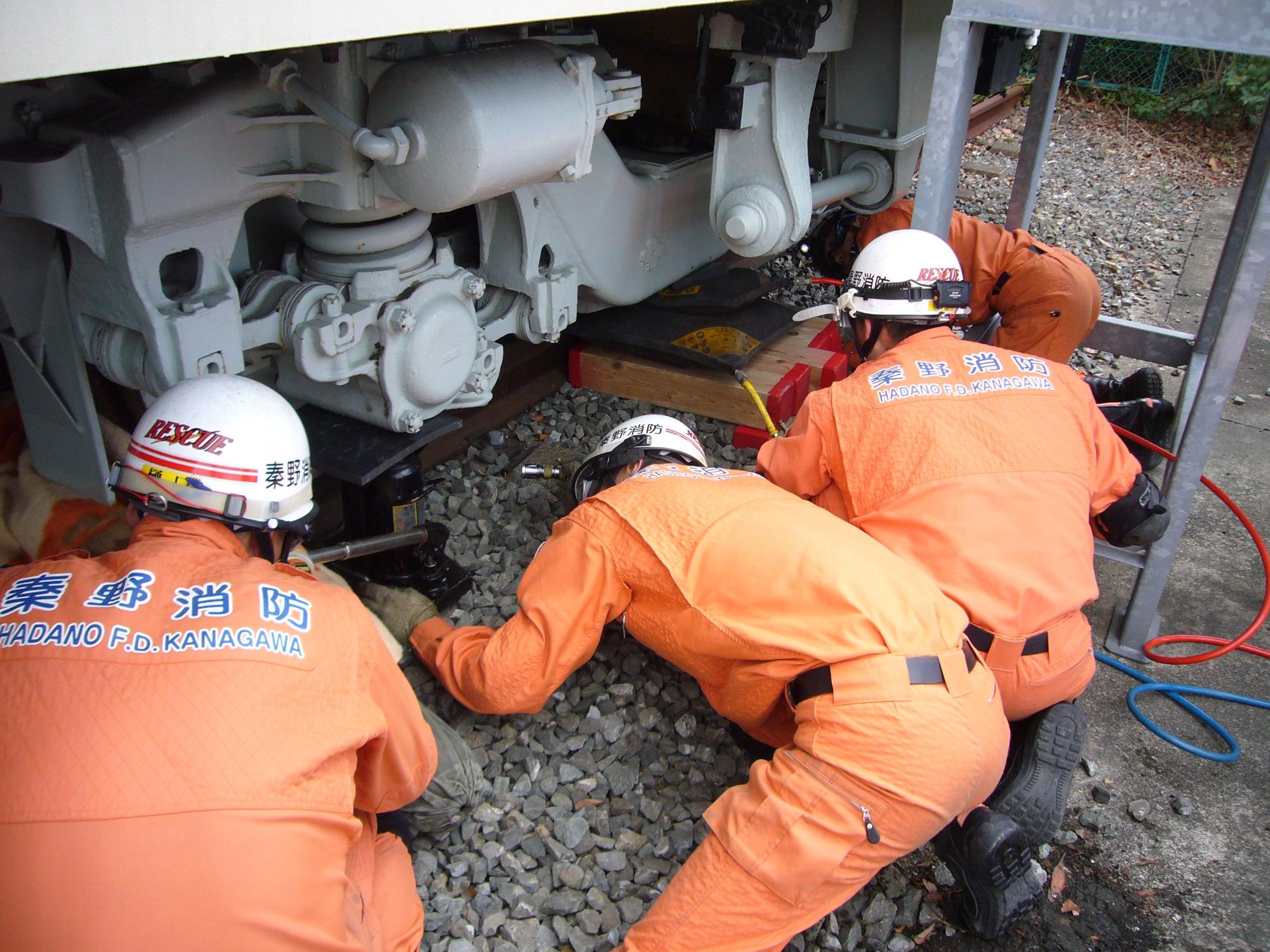 電車の台車下部で、オレンジ色の活動服と白ヘルメットを着用した3人の救助隊員が、線路の砂利の上に身をかがめながら救助活動を行う訓練風景の写真