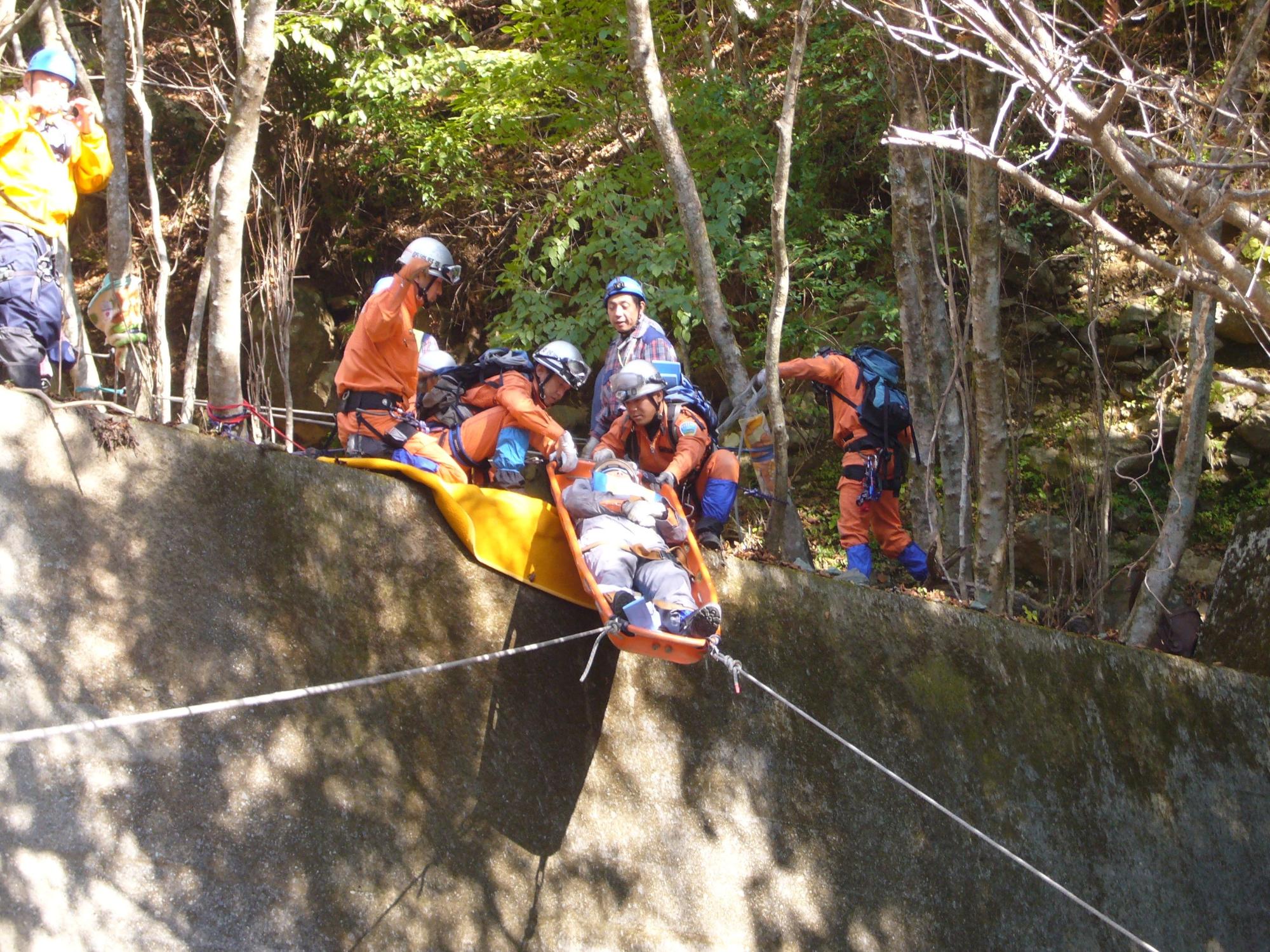 自然林の中のコンクリートの壁の上で、オレンジ色の活動服を着た複数の隊員が、要救助者に見立てた人物をストレッチャーに乗せてロープで吊り上げる山岳救助訓練風景の写真