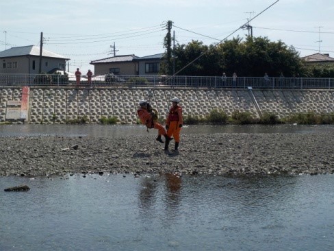 浅い川の砂利地帯で、オレンジ色の活動服を着た救助隊員がワイヤーで吊るされた要救助者に見立てた人形を受け取る河川救助訓練の風景を、対岸の家屋と電線とともに捉えた写真