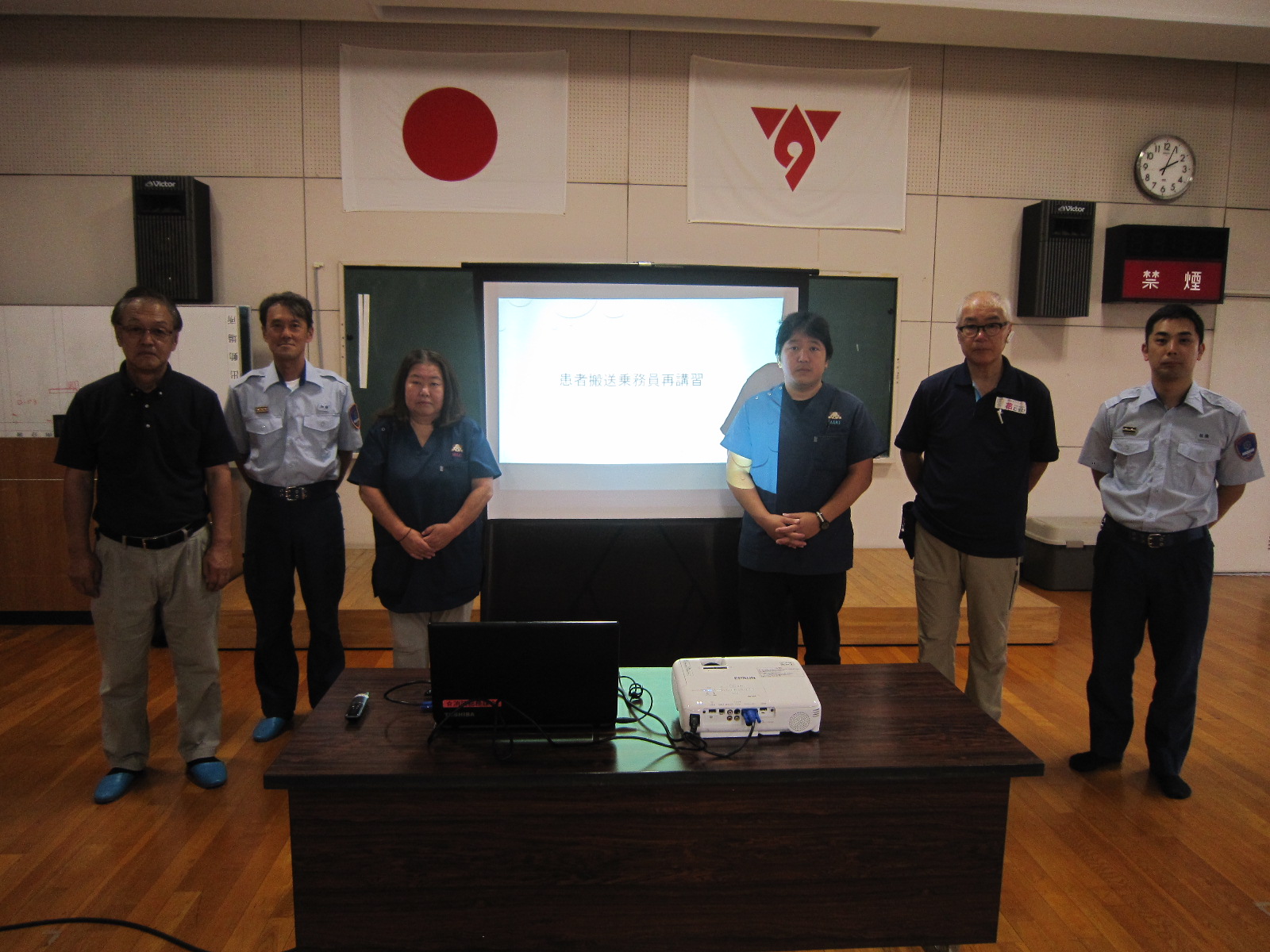 「患者搬送乗務員再講習」と映るスクリーンの前に、制服や医療用ウェアを着用した男女6人が横一列に並んで立っている集合写真