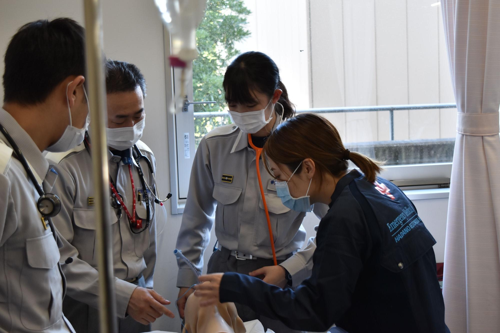 室内で、制服とマスクを着用した4人の救急隊員が、訓練用人形を囲み、一人の女性が処置を施しながら他の3人が真剣に見守っている様子の写真