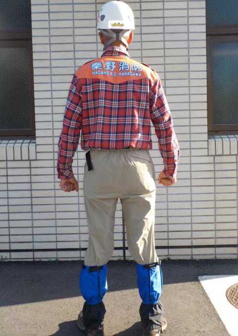 赤いチェックのシャツの背中の肩甲骨付近に「秦野消防 HADANO F.D. KANAGAWA」と青い文字で書かれたオレンジ色の布が縫い付けられ、ベージュのパンツと青いスパッツ、白いヘルメットを着用し、登山靴を履いてレンガ状の壁を背に立つ、救助隊員を後ろから撮影した写真