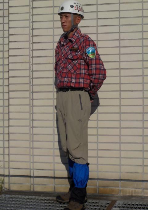 白いクライミングヘルメットを着用し、赤と黒のチェックのシャツの左袖に山岳救助隊のワッペンをつけ、ベージュのパンツと青いスパッツ、登山靴を履いてレンガ状の壁を背に立つ、救助隊員を斜め前より撮影した写真