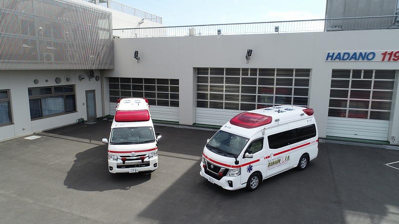 「HADANO 119」と書かれた消防署の建物の車庫前にある舗装された広場に、白い車体に赤いラインが入った2台の救急車が斜めに停まっている写真