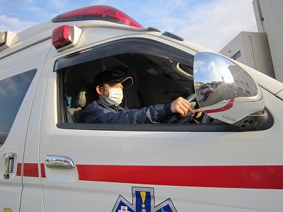 白い車体に赤いラインが入った救急車に、青い制服と黒い帽子、白いマスクを着用した救急隊員が、まぶしそうにハンドルを握っている写真