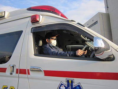 白い車体に赤いラインが入った救急車の運転席で、青い制服と黒い帽子、白いマスクを着用した救急隊員がサングラスを着用している写真