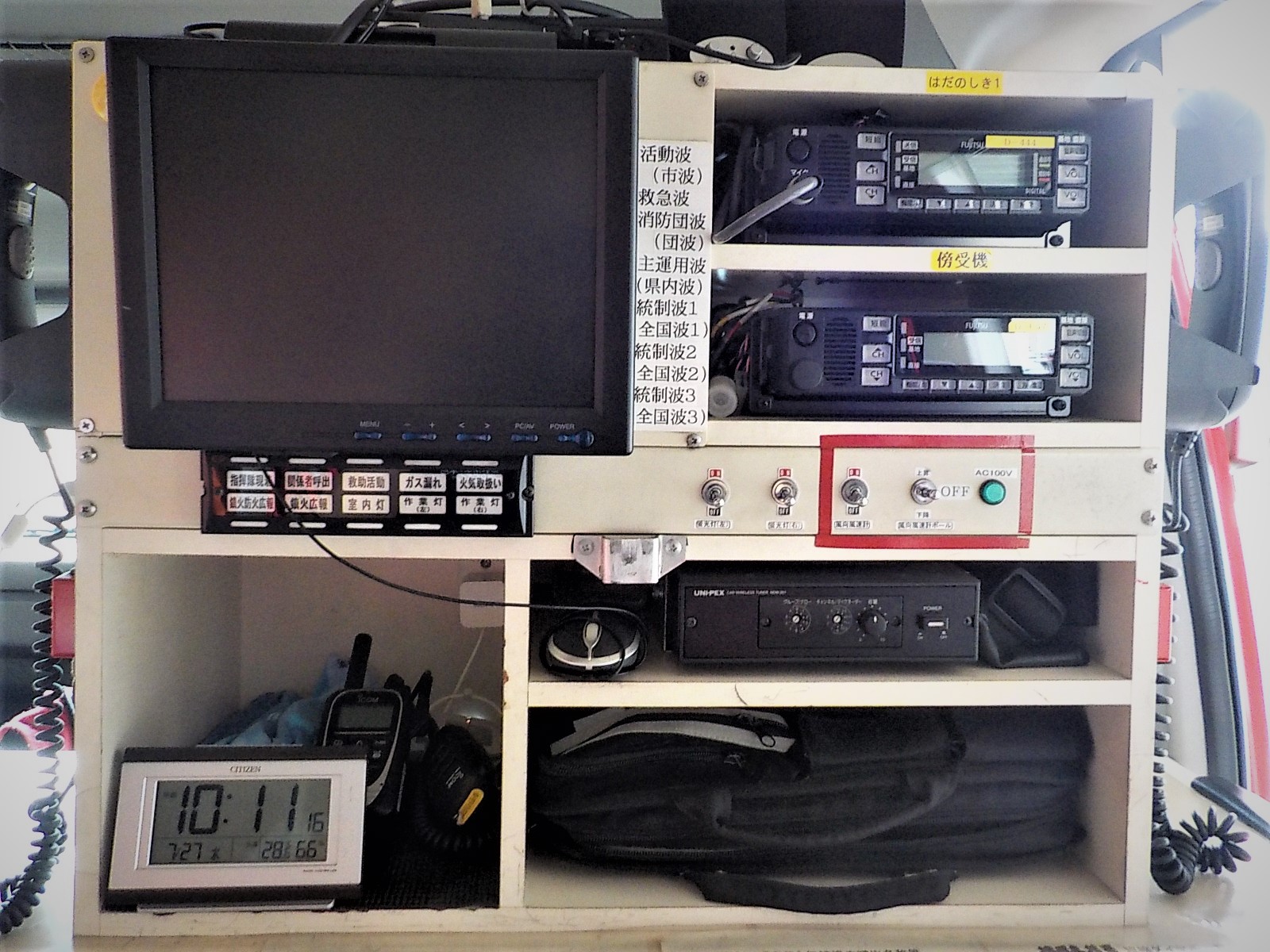 消防車両の内部設置された棚のアップで、液晶モニター、複数の無線機、電源盤、時計、トランシーバーなどの通信・情報処理機器が整理され並んでいる写真