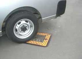 シルバーの自動車のリアタイヤが、アスファルトの上に設置された黒とオレンジの模様が入った消防水利を踏みつけている様子の写真