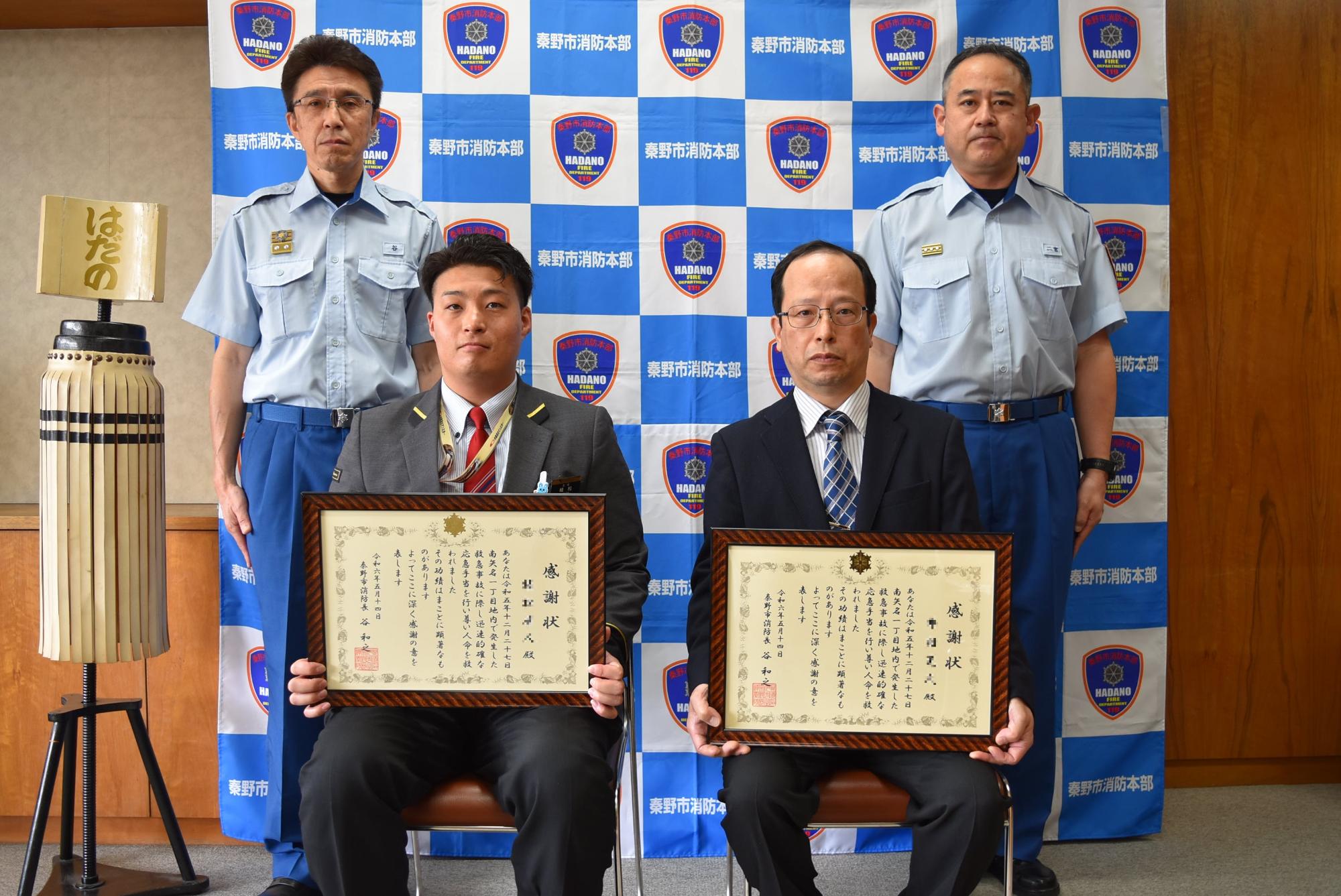 秦野市消防本部の背景前で、表彰状を手にした2人が椅子に座り、制服姿の消防署職員2人が背後に立った記念写真