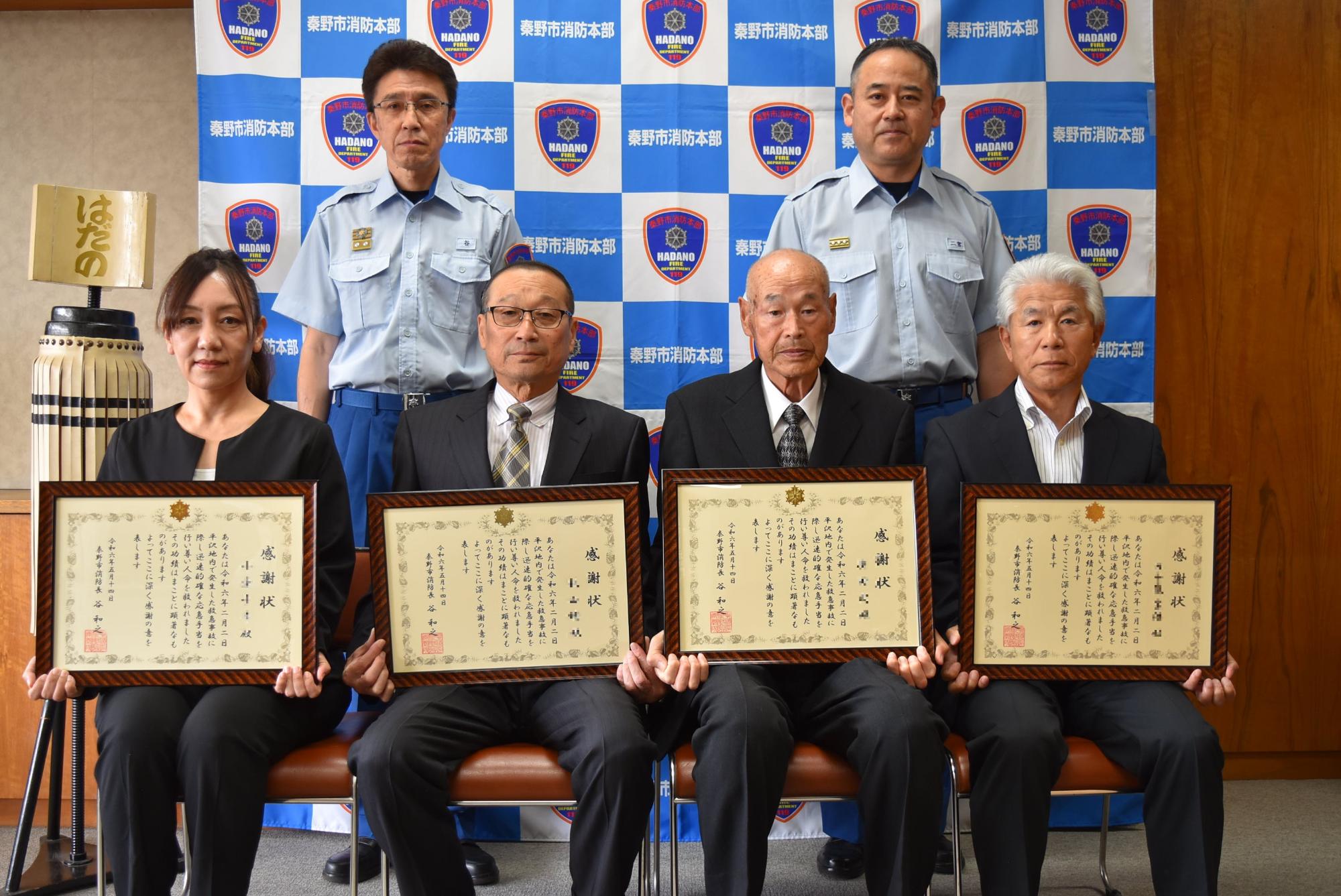 秦野市消防本部のロゴが並ぶ背景の前で、後方に立つ制服姿の消防署職員の男性2人の前で、それぞれ感謝状を手にしたスーツ姿の男性3人と女性1人が並んで座った記念写真