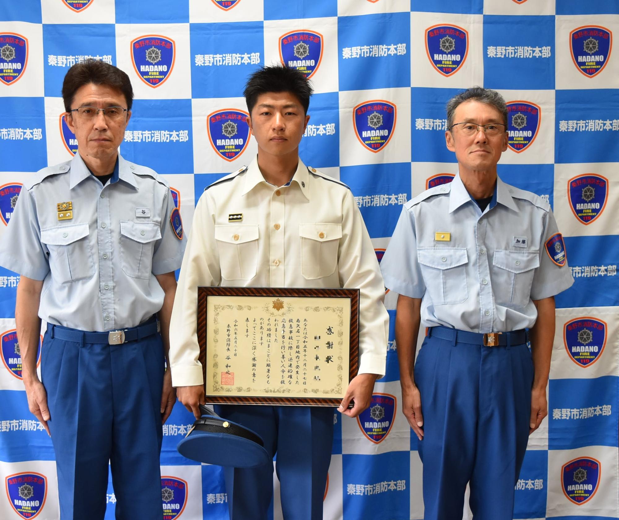 秦野市消防本部のロゴが並ぶ青い背景の前で、中央に立つ白い制服の男性が賞状を手に持ち、左右の水色の制服を着た消防署職員の男性2人と共に写した記念写真