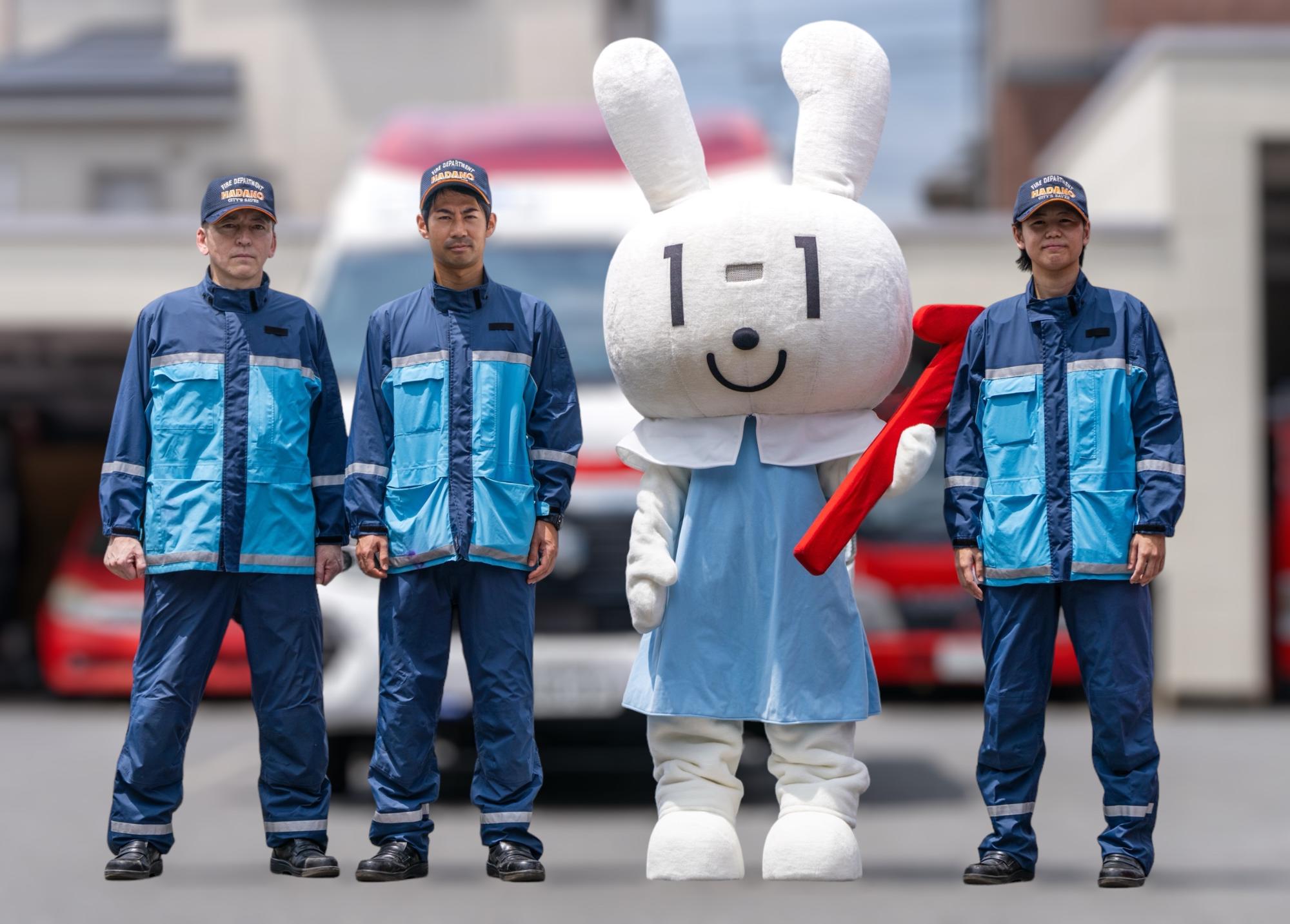 救急車を背に、青と水色の活動服を着た3人の救急隊と、赤いバトンを持っているマイナンバーPRキャラクターの「マイナちゃん」が並んで立っている写真