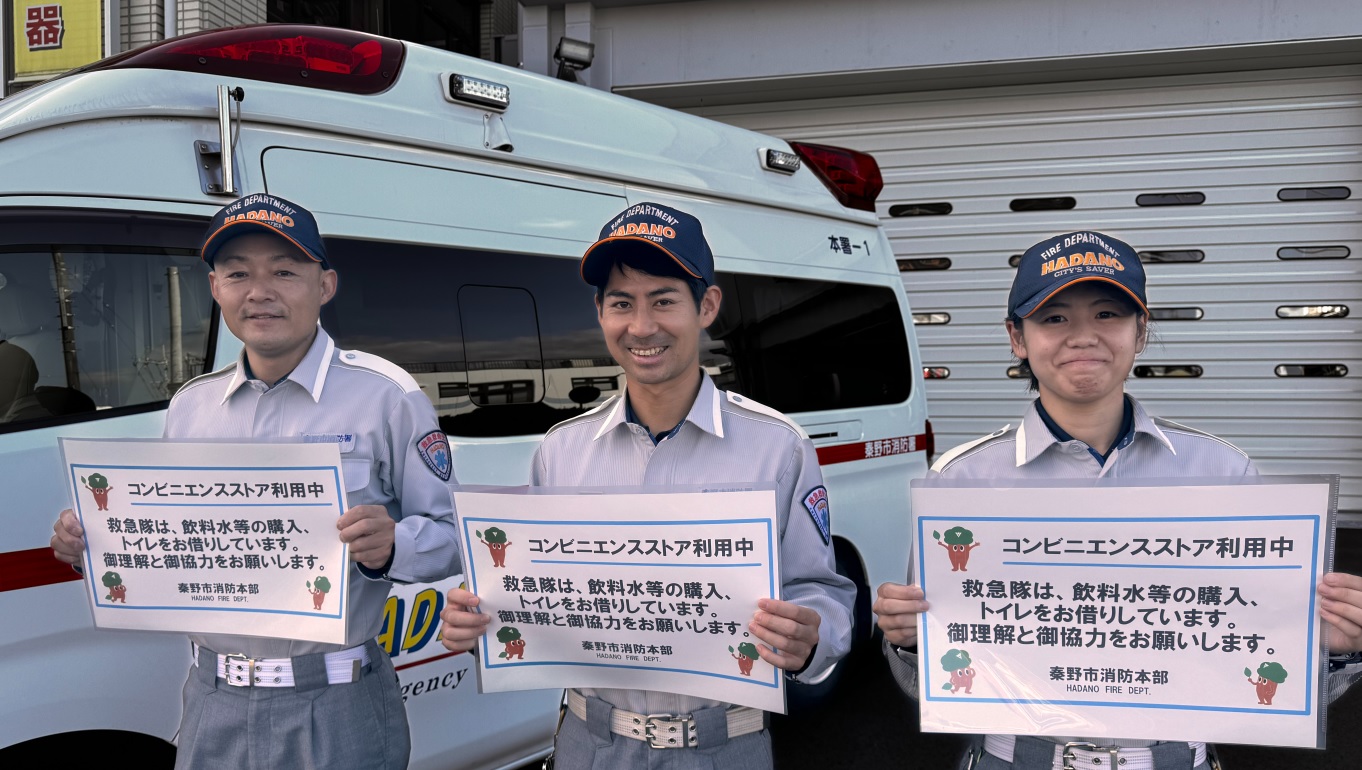 制服姿の秦野市消防本部の隊員3名が救急車を背に、「コンビニエンスストア利用中 救急隊は、飲料水等の購入、トイレをお借りしています。御理解と御協力をお願いします。」と書かれた紙を笑顔で掲げている写真