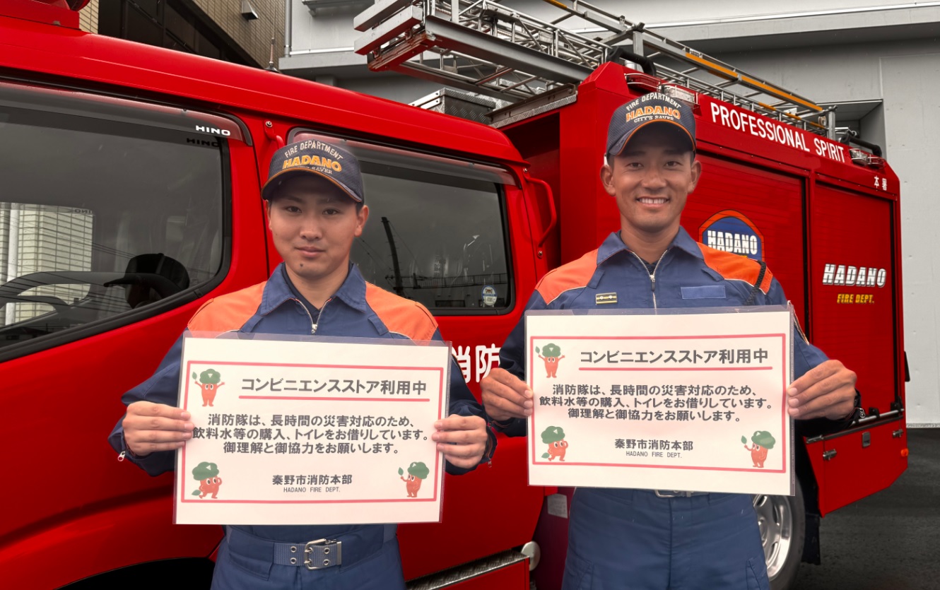 消防車を背にした秦野市消防本部の隊員2名が、「コンビニエンスストア利用中 消防隊は、長時間の災害対応のため、飲料水等の購入、トイレをお借りしています。御理解と御協力をお願いします。」というコンビニ利用への理解を求める案内を笑顔で掲げている写真