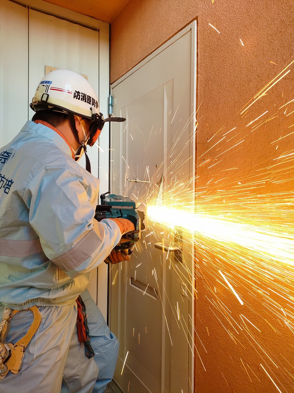 防塵マスクとヘルメットを装着した救助隊員が、電動カッターを使用して白い金属製のドアの鍵付近を切断しており、その際に発生した大量の火花が激しく飛び散っている、救助訓練の様子の写真