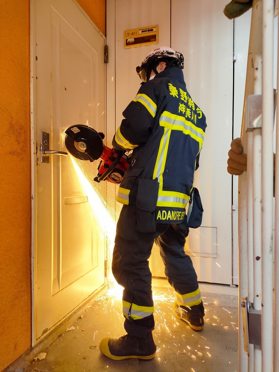 紺色の活動服と反射材を着用しヘルメットを被った救助隊員が、電動切断機を使って頑丈な白いドアの鍵部分を切断しており、その際に飛び散る激しい火花が周囲を明るく照らしている救助訓練の様子の写真
