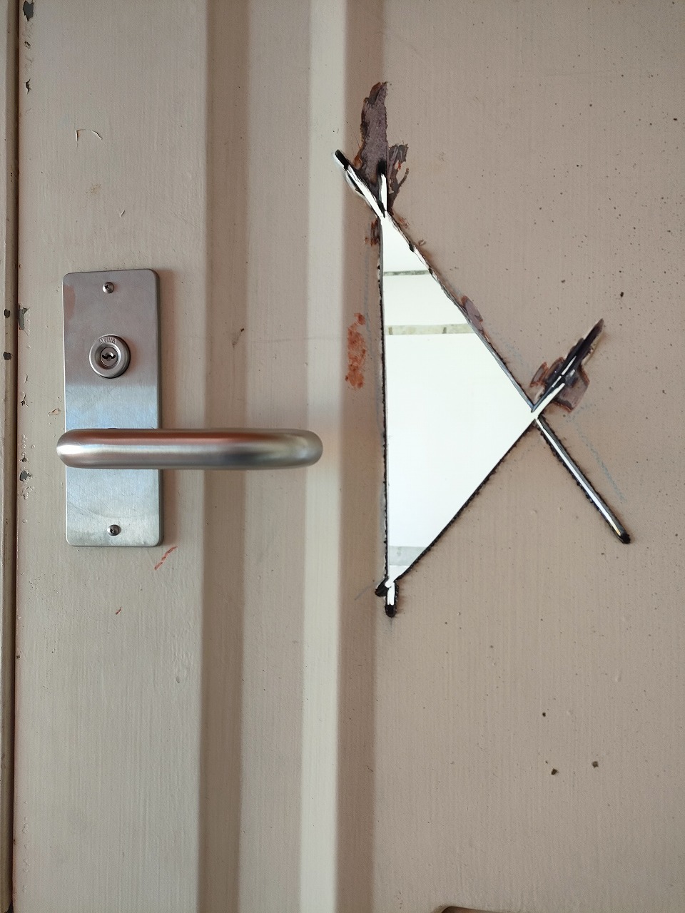 アイボリー色の金属製ドアの表面に、光沢のある銀色のレバーハンドル錠が取り付けられており、その右側には救助活動などで開けられたとみられる、破断面がギザギザとした三角形の穴が縦長に開いているクローズアップ写真