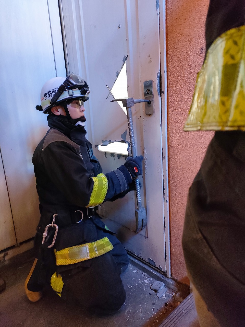 反射材付きの濃紺の救助服とヘルメットを着用した救助隊員が、破られた白い金属製ドアの錠前付近にバールのような工具を差し込み、膝立ちで破壊活動を行っている救助訓練の様子の写真