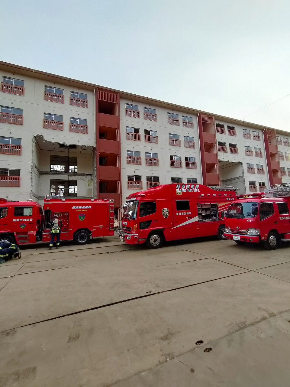 4台の赤い消防車両が並ぶ広場に面して、壁の一部が破壊された5階建てのベージュと茶色の集合住宅がそびえ立ち、倒壊建物からの救助活動訓練が行われている様子の写真