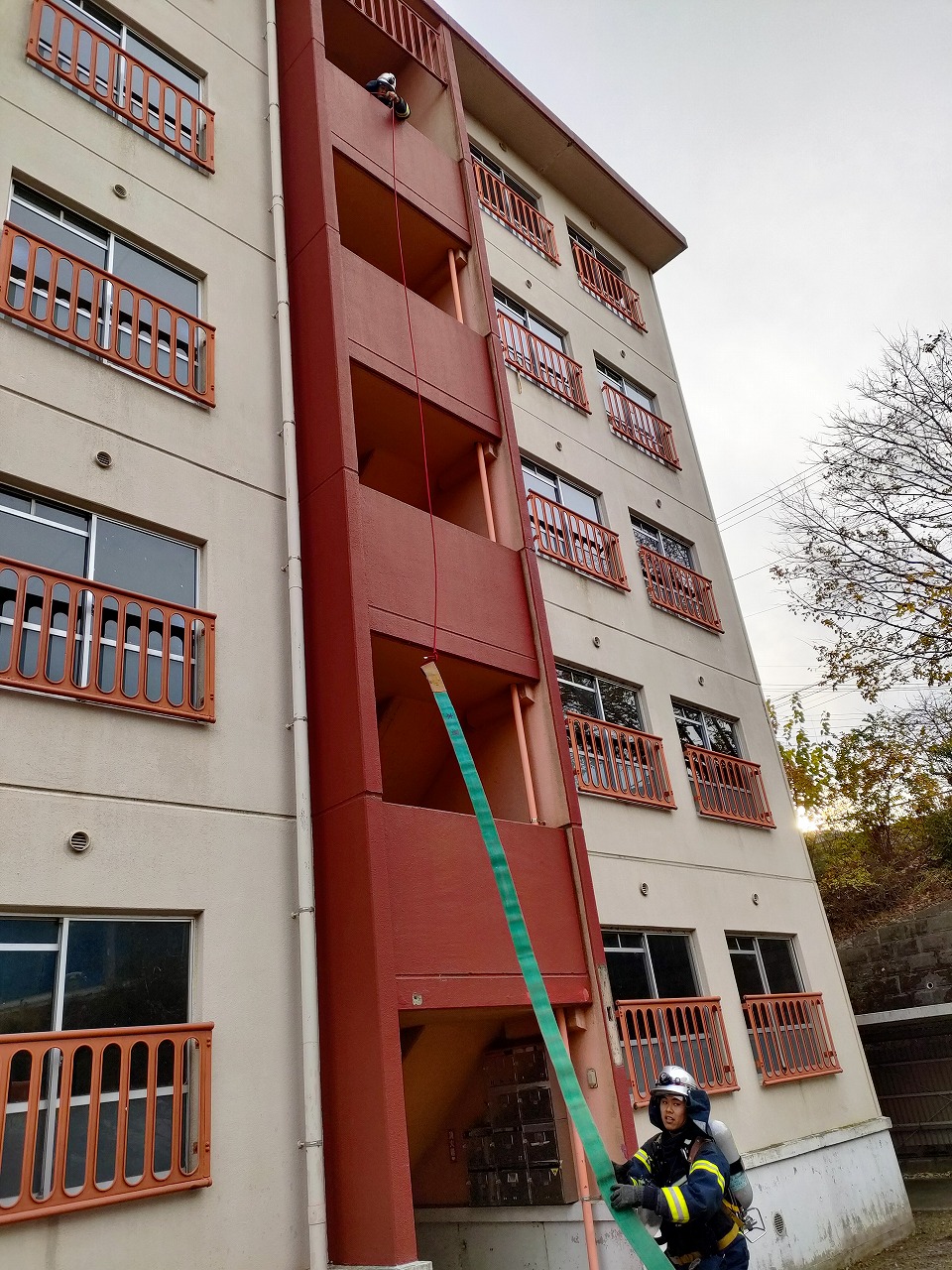 中層の集合住宅の外壁で、ヘルメットと酸素ボンベを背負った救助隊員が建物の屋上から降ろされたロープと緑色のホースを操作し、人命救助訓練を行っている様子を下から見上げる構図で撮影した写真