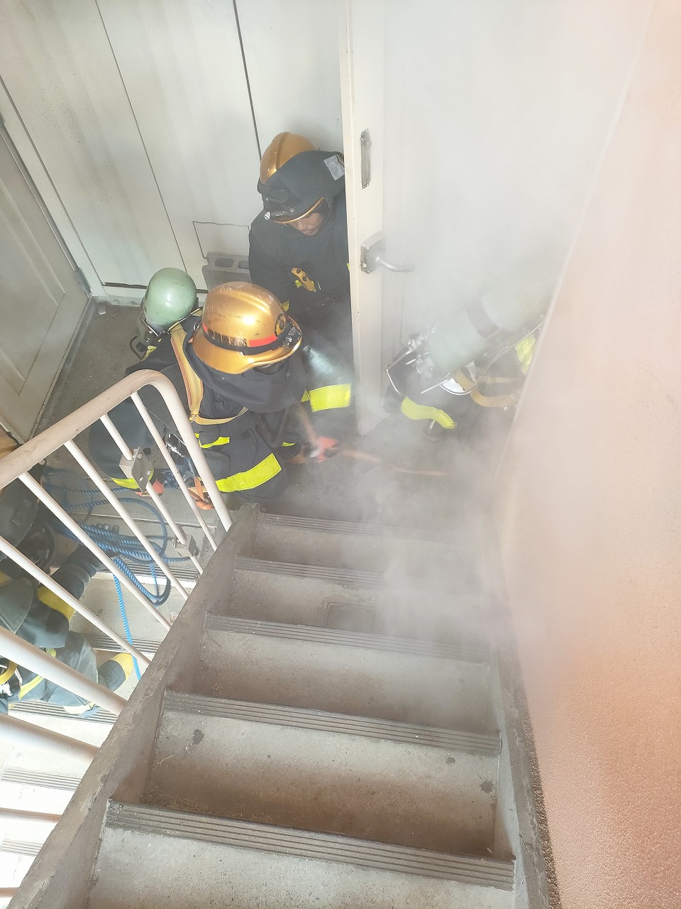 建物内部の階段上り口付近で、ヘルメットや防火服を着用した複数の消防隊員が、煙が立ち込める中、ドアを開けて建物内部へ侵入しようとしている緊迫した訓練の様子の写真