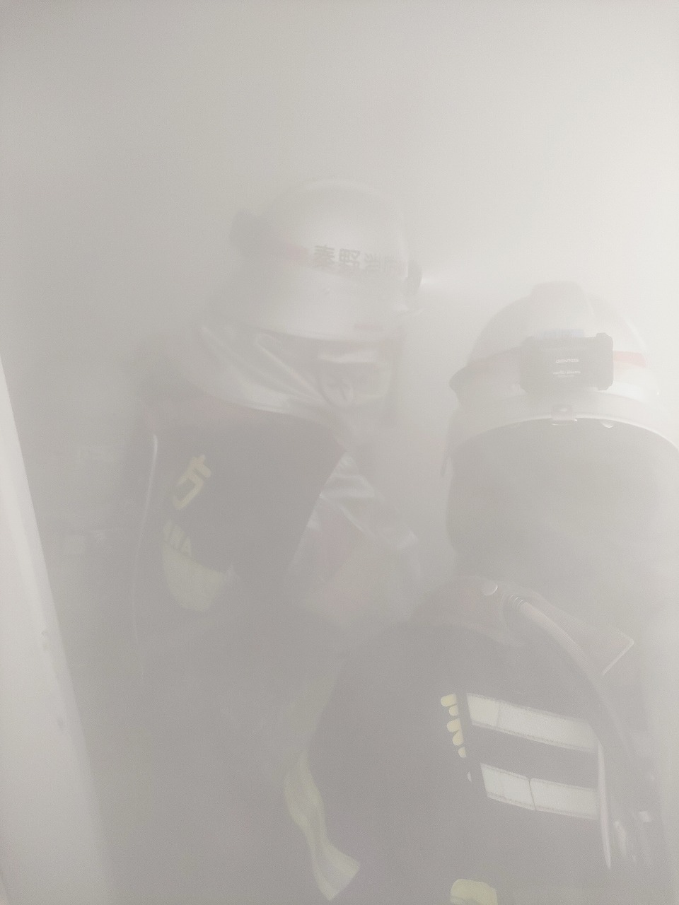 ヘルメットと防火服を着用した2名の消防隊員が、顔や体がほとんど見えないほどの濃い白い煙や霧が充満する建物内部で、視界の悪い極めて困難な状況下での活動を行っている救助訓練の様子の写真