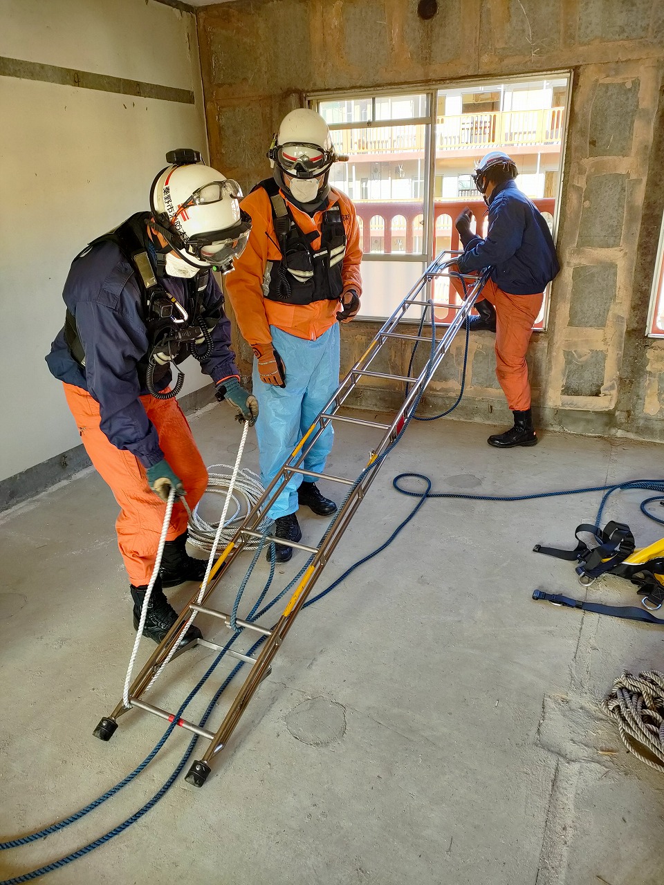 レスキュー隊員3名が、防塵マスク付きのヘルメットとオレンジ色のズボンを着用し、建物内部の窓際で、ロープを組んだ金属製の長いはしごを使った救助訓練を行っている様子の写真