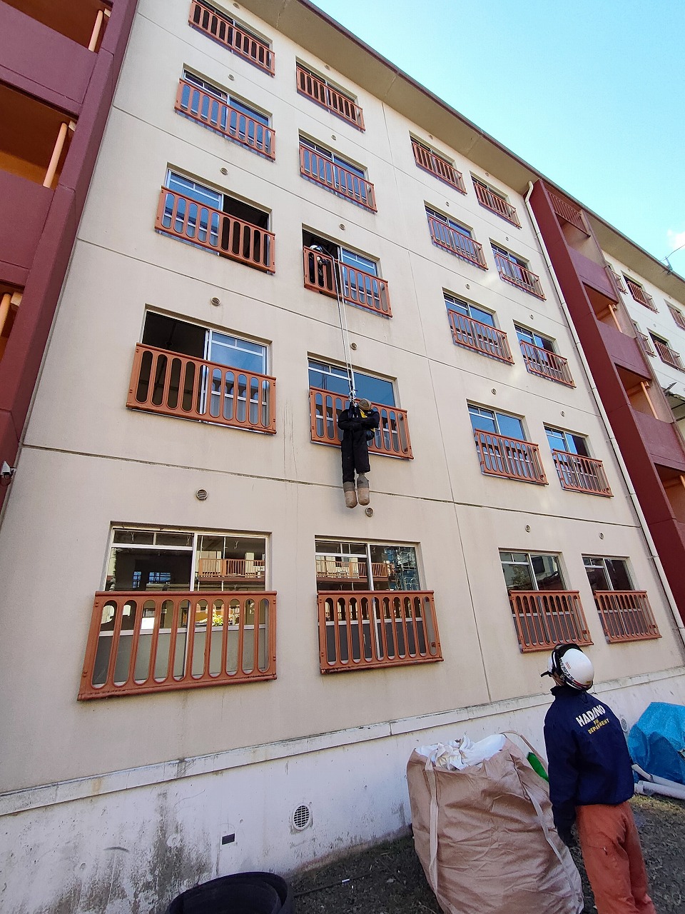 集合住宅の外壁で、ヘルメットを装着した救助隊員が、上の階の窓から吊り下げられたロープを使って懸垂下降している様子を、地上から別の隊員が見上げている訓練風景の写真