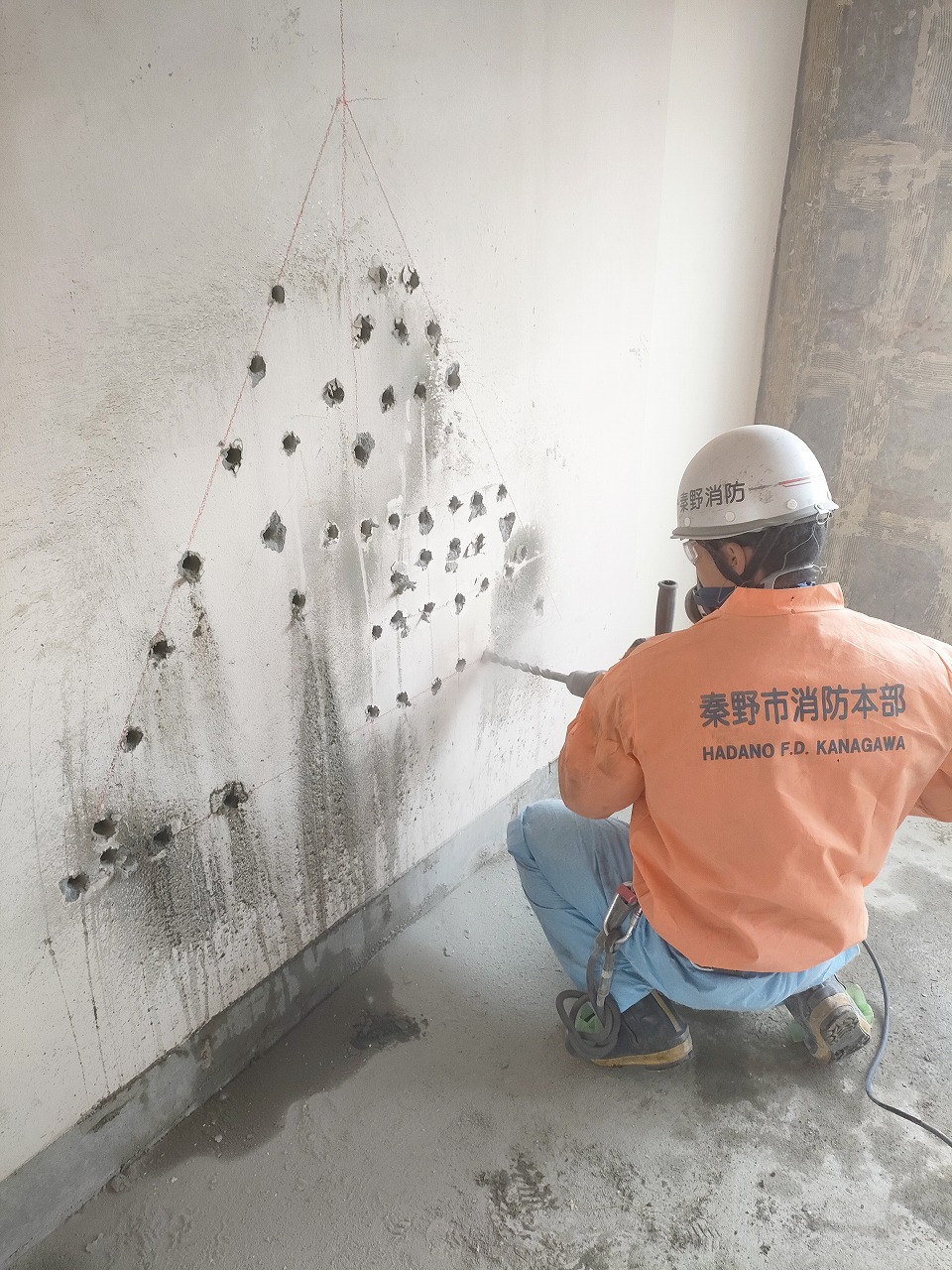 ヘルメットにマスク姿の「秦野市消防本部」のオレンジ色の作業服を着た男性が、コンクリートの壁にドリルで無数の穴を三角形に穿孔している、訓練の様子の写真