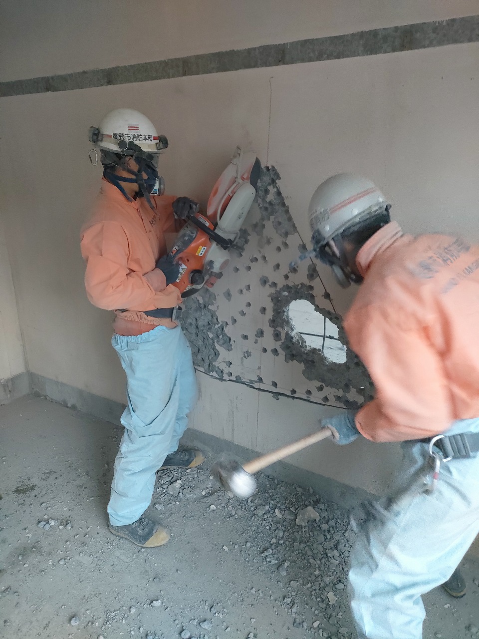 マスクとヘルメットを装着した2名の作業員が、オレンジ色の作業服と水色のズボンを着用し、1人はエンジンカッターでもう一人はハンマーで壁のコンクリートを破砕している、訓練の作業風景の写真