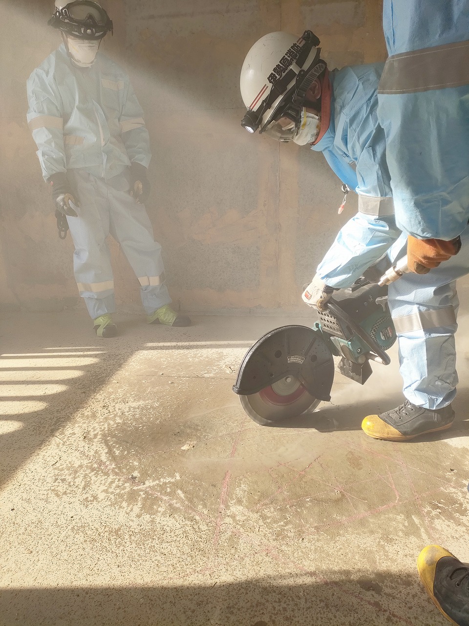 青い防護服とヘルメットを着用した救助隊員が、濃い粉塵の中で、切断機を使ってコンクリート床を切断する訓練を行っており、別の隊員がその様子を煙の中に立って見守っている作業風景の写真