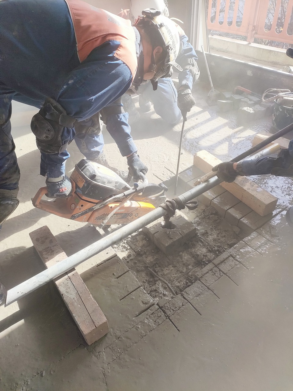 青い作業服を着た作業員が、コンクリートの床に載せたオレンジ色のエンジンカッターを操作し、水と泥が飛び散る中で、足元のパイプやレンガをガイドとして床を切断している建築現場の様子の写真