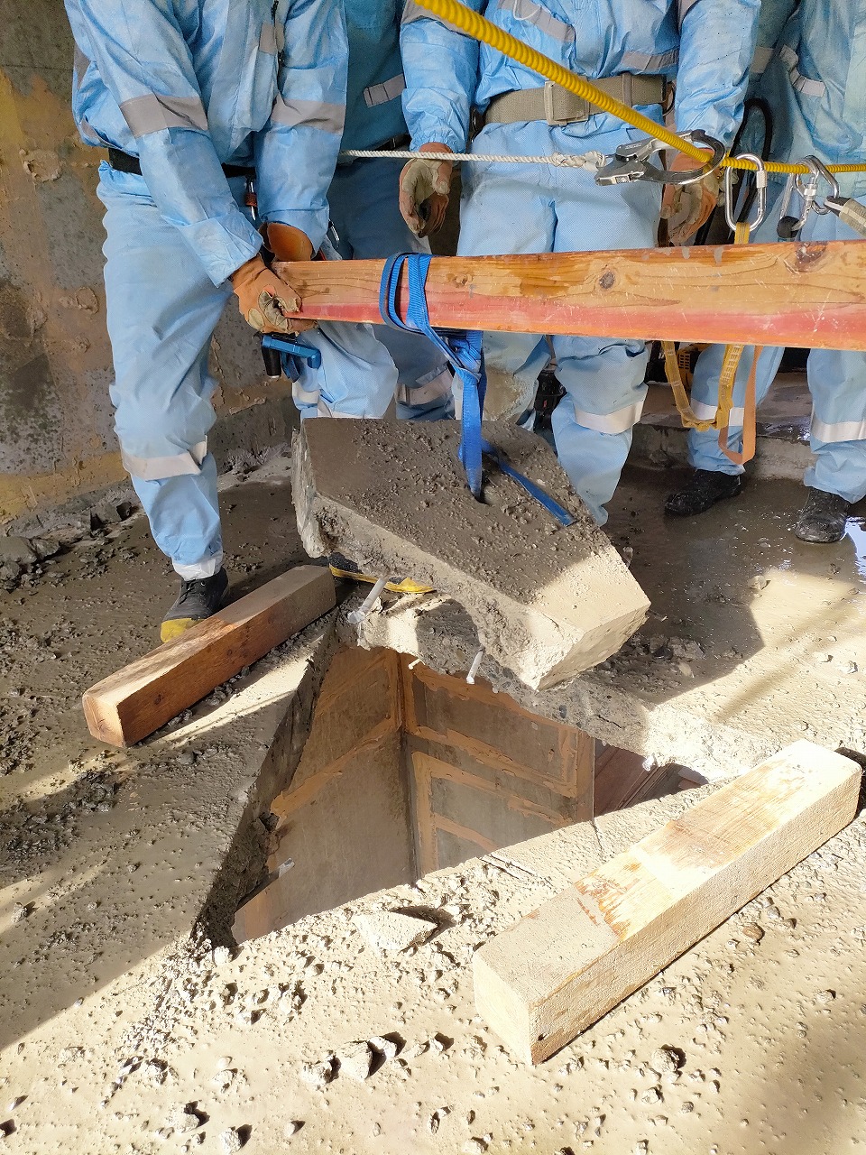 数名の作業員が明るい青色の作業服を着用し、オレンジ色の木材と青いスリングを使って、掘削された穴から大きなコンクリート片を慎重に吊り上げて運び出している建設作業の様子の写真