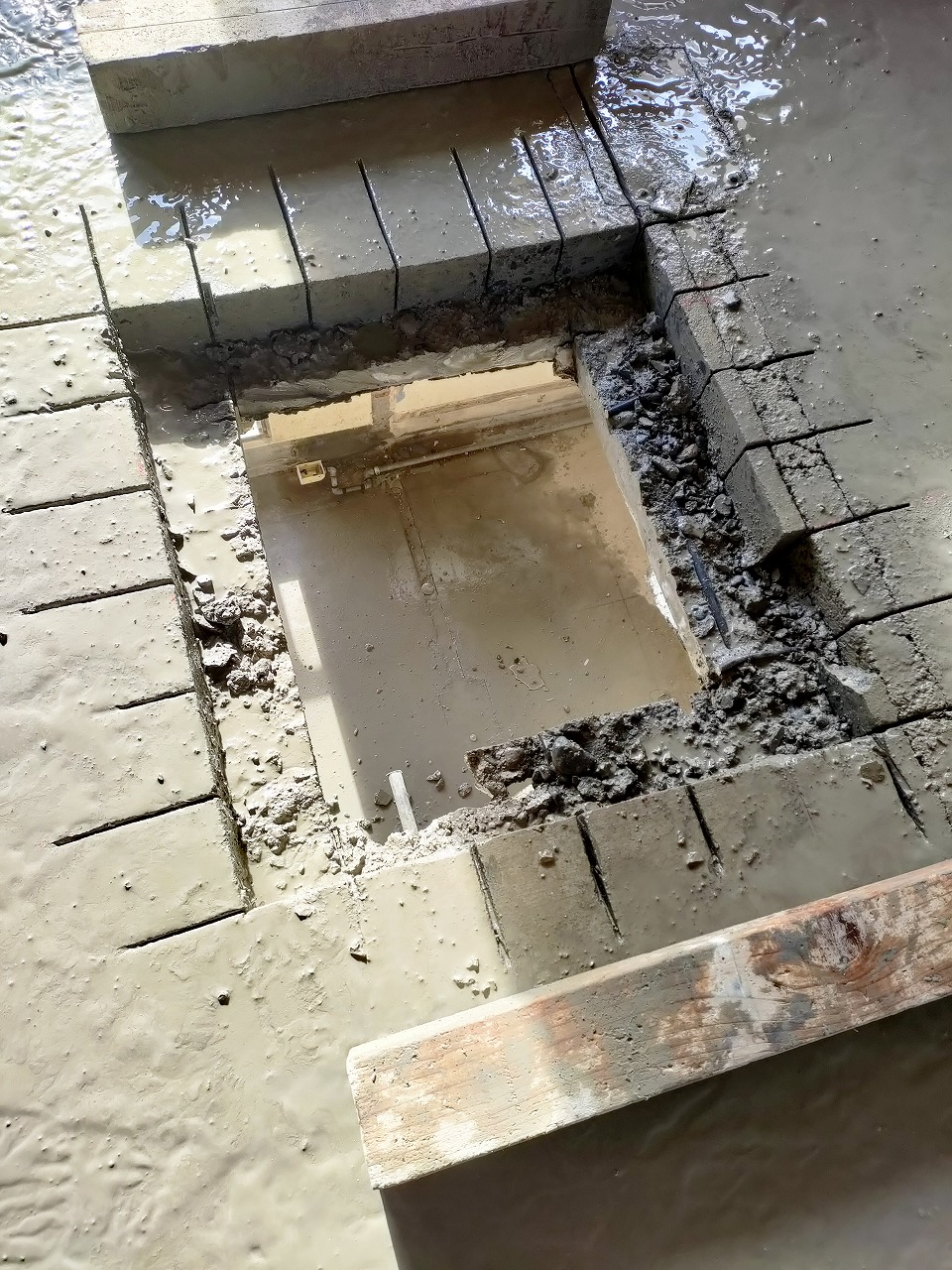 セメントのような泥水で覆われた床の一部を四角く切断し、周囲には切断用の溝が細かく入れられたコンクリート片が並べられ、その奥に深い穴が開いている建設現場の様子の写真