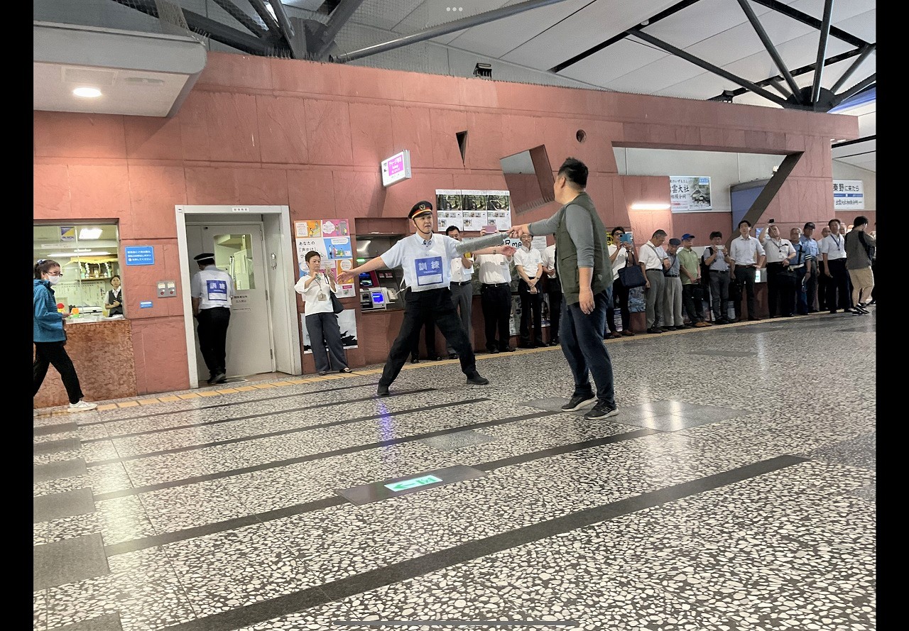 タイル張りの駅構内で、多くの人が周囲で見守っている中、白い制服と帽子を着用した駅員らしき人物が両腕を広げて正面の男性を制止しているような様子を捉えた写真