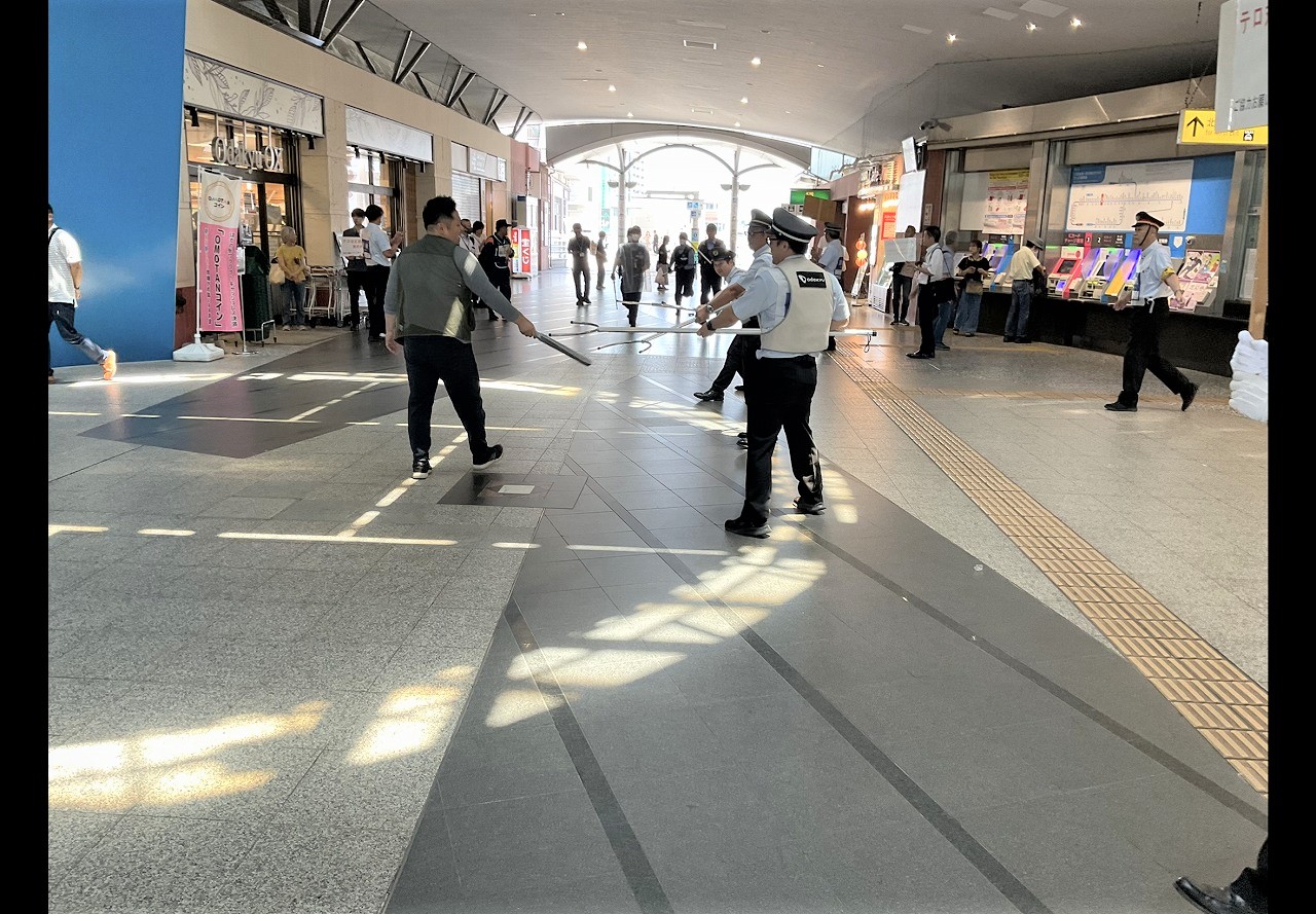 広い駅構内通路で周囲には多くの人が立って様子を見ている中、警察官や駅員らしき制服姿の二人が長い棒状のものを持って進行方向の男性に対応している写真