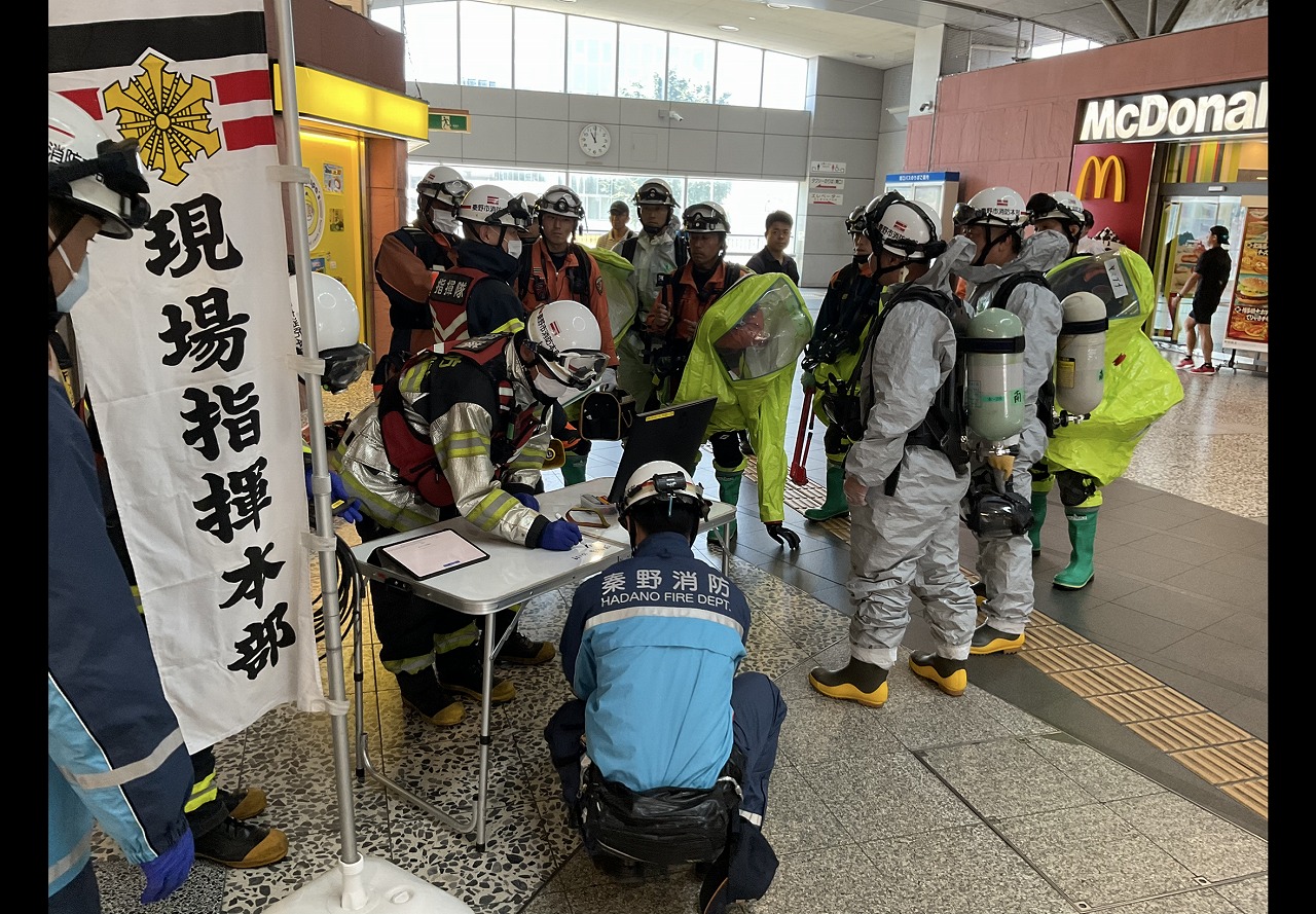 駅構内に「現場指揮本部」と書かれたのぼりが立てられ、秦野消防の青い服を着た隊員を中心に、化学防護服やオレンジ色の活動服を着用しヘルメットをかぶった複数の隊員が、情報共有のために集まり小さなテーブルで記録を確認している訓練の写真